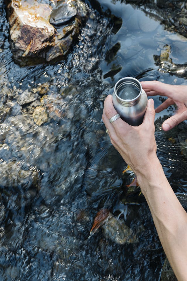 Hand holding a cup of water beside river rocks, symbolizing clarity and refreshment.